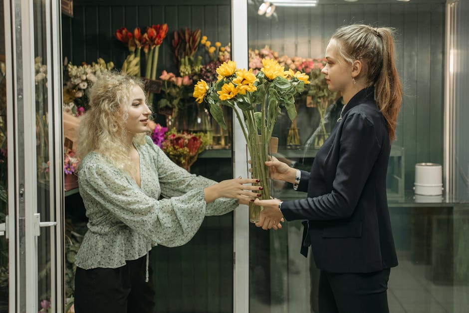 A florist with wavy blonde hair, wearing a light green, patterned blouse, is handing a bouquet of bright yellow sunflowers wrapped in clear plastic to a customer with long brown hair tied back in a hi