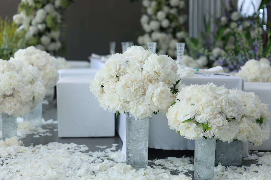 Elegant floral arrangements featuring large bouquets of white peonies and hydrangeas displayed in modern, textured concrete vases atop a table covered with white petals, set in a well-lit indoor venue