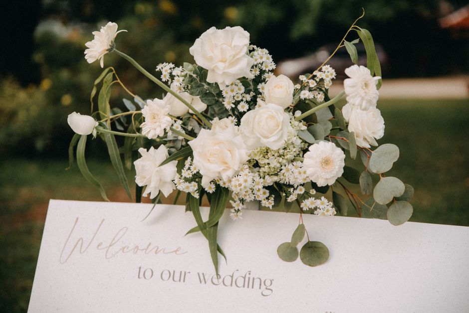 A delicate bouquet of fresh white flowers including roses, peonies, daisies, and small blossoms, with green eucalyptus and leafy accents, arranged in a natural, slightly asymmetrical style. The flower