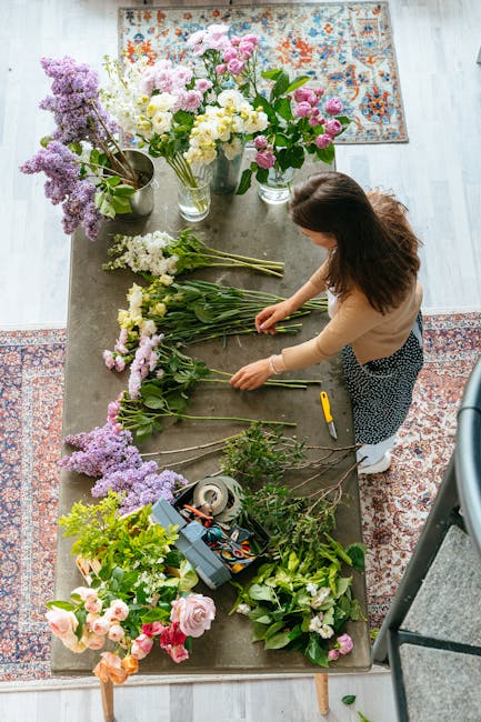 A woman with brown hair, dressed in a beige top and patterned skirt, arranging a variety of fresh flowers on a rustic wooden table in a well-lit room. The table is covered with an assortment of flower