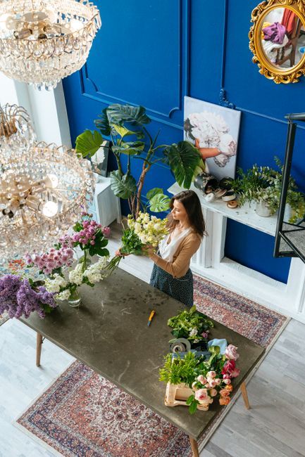A woman arranging a floral bouquet inside a bright, modern florist shop with vivid blue walls. The arrangement includes white and pink flowers, with large green tropical leaves adding volume and textu