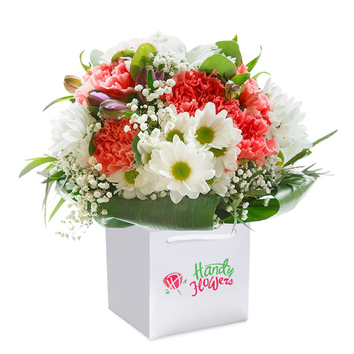 Expertly arranged by a professional florist in the heart of Sipson, this abundant bouquet stands tall in a crisp, contemporary white gift bag, its clean lines and solidity suggesting readiness to grace any home or celebration. The display is one of vivid colour and graceful harmony: plush coral and light peach carnations form the core, each petal a testament to nature's artistry. Scattered amongst them, pristine white daisies with vibrant yellow-green centers catch the light, exuding a refreshing charm reminiscent of the summer mornings near Sipson's well-loved pathways. Airy clusters of white gypsophila emerge between the main blossoms, infusing the arrangement with delicacy while adding fullness that softens the bordered structure. Nestled discreetly are a few tightly closed buds, veiled in hints of purple and suggesting new growth and unfolding stories. Surrounding the bouquet, glossy deep green foliage-broad and lush-forms a natural collar, framing the flowers and providing an elegant contrast to the pastel hues above. The entire composition is illuminated by gentle, diffuse lighting, which enhances the richness of every colour and texture. Sturdy ribbon handles above the bag suggest convenience and an effortless delivery experience for local Sipson recipients. This meticulously crafted bouquet, with its rounded, generous shape and flawless freshness, speaks of joyful moments and sincere sentiment-a gift suited for birthdays, anniversaries, or simply to bring delight to someone special within the Sipson community.